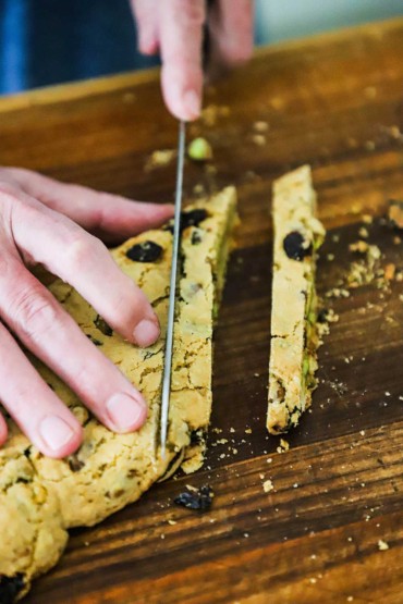 A person using a serrated knife to cut a baked loaf of biscotti into thin strips on a cutting board.