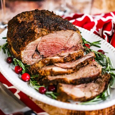 A close-up view of a roasted boneless leg of lamb with garlic and herbs sitting on white platter garnished with rosemary sprigs and cranberries.