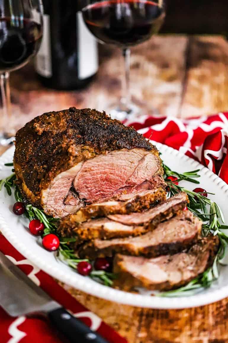 A close-up view of a roasted boneless leg of lamb with garlic and herbs sitting on a white platter garnished with rosemary sprigs and cranberries.