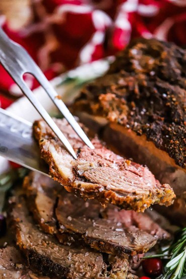 A meat carving meat fork and knife being used to lift a slice of boneless leg of lamb from the roast on a platter.