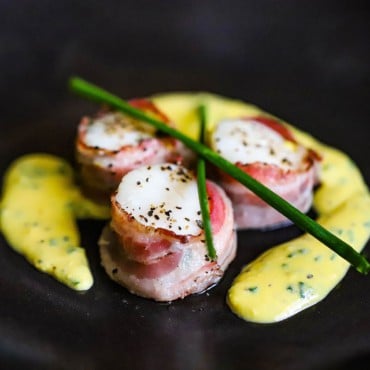 A close-up view of sea scallops with pancetta wrapped around them sitting on a black plate with an egg and chive sauce surrounding them.