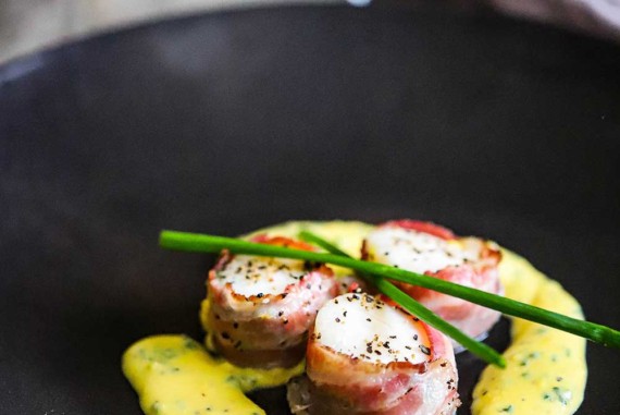 Three sea scallops with pancetta sitting on a black plate with a yellow egg and chive sauce on the plate, as well.