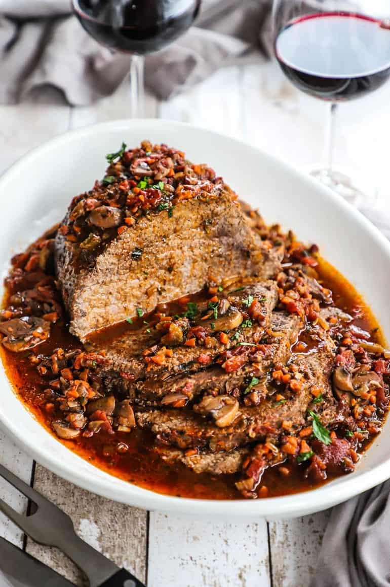 An Italian pot roast that has been sliced and is sitting in a large oval platter with a tomato and mushroom sauce all over it.