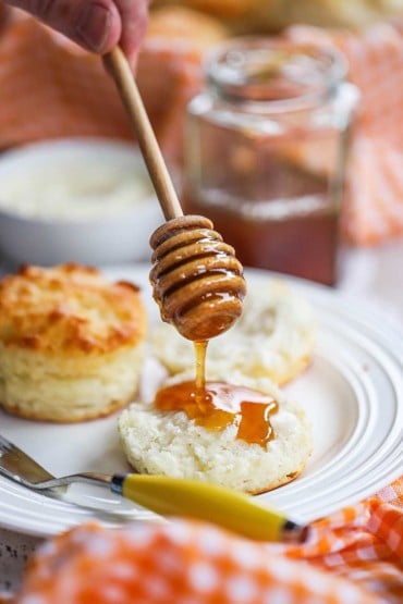 One drizzles golden honey with a honey stick over cracked Southern biscuits on a plate.