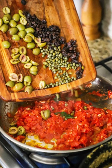 A cutting board being used to transfer olives, capes, and raisins into a skillet of cooked diced tomatoes.