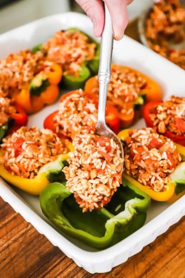 A person using a large silver spoon to transfer a ground turkey and rice filling into the cavity of an open green bell pepper in a dish.