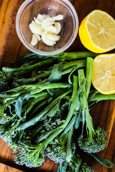 An overhead view of fresh broccolini next to a small bowl of garlic cloves and a lemon that has been halved.
