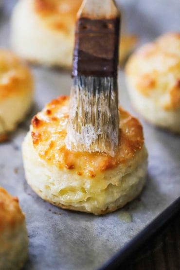 A pastry brush being used to smear melted butter over the top of a Southern biscuit on parchment paper.