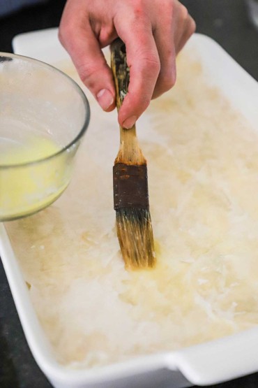 A person using a pastry brush to apply melted butter onto a sheet phyllo dough that is sitting in a baking dish.