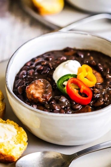 A close-up view of a soup bowl filled with black bean and andouille sausage topped with sliced peppers and a dollop of sour cream on a plate with a piece of cornbread on it.