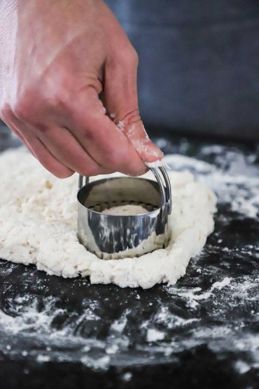 A person using a biscuit cutter to cut a round piece of dough from a slab of biscuit dough on a black marble counter.