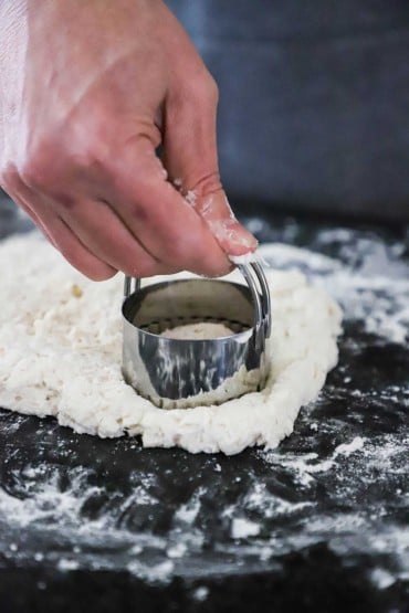 A person cuts a round piece of dough with a cookie cutter from a piece of cookie dough on a black marble counter.