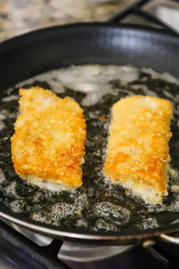 Two Parmesan crusted cod filets that are being lightly fried in a non-stick skillet with bubbling oil.