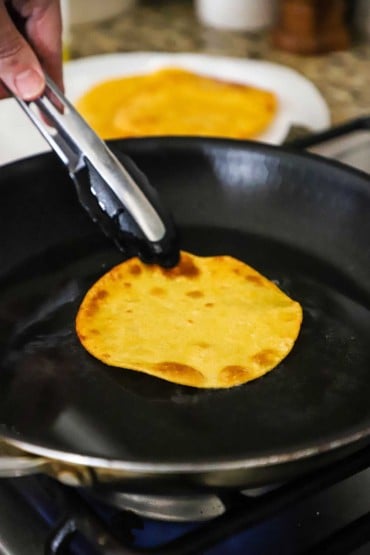 A corn tortilla being lifted up with a pair of tongs and is being lightly fried in a large non-stick skillet.