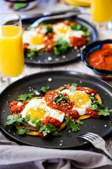 Two black plates filled with a serving of huevos rancheros both with a glass of orange juice nearby.