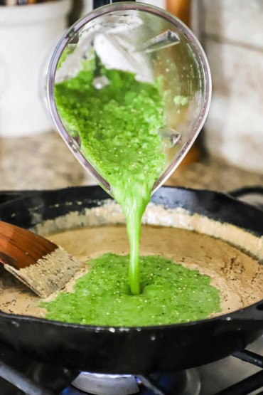A bright green tomatillo sauce being poured from a blender into a skillet filled with a puréed pumpkin sauce.