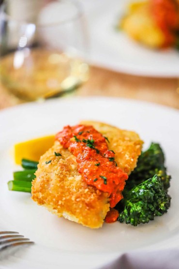A white dinner plate filled with Parmesan crusted cod with romesco resting on sautéd broccolini.