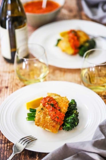 Two dinner plated filled with a serving of parmesan crusted cod with romesco and sautéd broccolini next to two glasses of white wine.