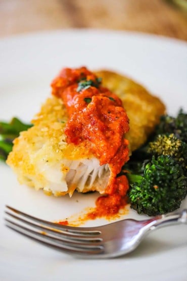 A close-up view of Parmesan crusted cod topped with romesco sauce and a bite of it is gone.