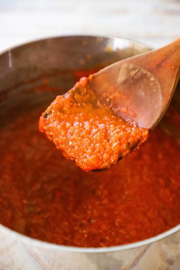 A wooden spoon holding ranchero sauce is being held up over a pan of the sauce.