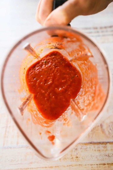 An overhead view of a blender holding puréed red bell peppers, tomatoes, and spices.