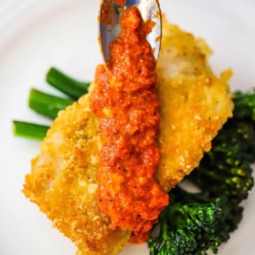 A small spoon being used to add romesco sauce over the top of Parmesan-crusted cod on top of a bunch of sautéed broccolini.