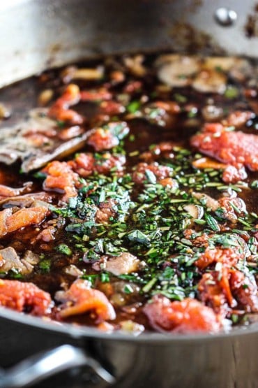 A large silver saucepan filled with a tomato and red wine sauce with fresh herbs that is simmering on the stove.