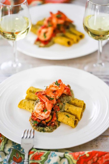 Two plates that contain a serving of Veracruz shrimp sitting on three rolled tortillas with pumpkin seed sauce and two glasses of white wine nearby.