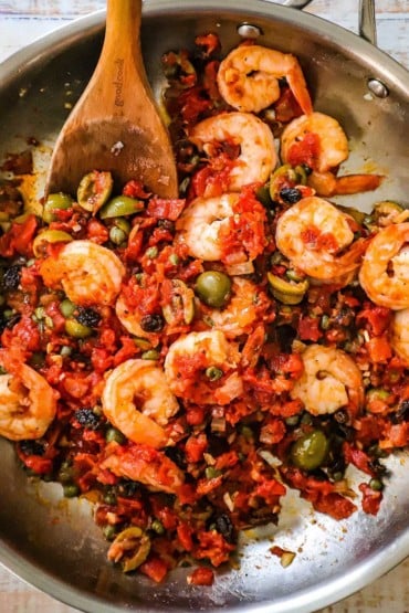 An overhead view of a stainless steel skillet filled with shrimp Veracruz with a wooden spatula stuck in the middle. 