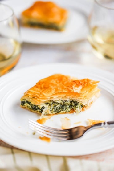 A square piece of spanakopita sitting on a white plate with a bite taken out of it.