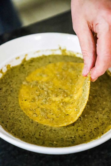 A person submerging a warmed corn tortilla into a bowl filled with pumpkin seed pipián sauce.