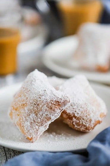 Two beignets that are covered with powdered sugar sitting on a dessert plate in front of a glass mug of coffee.