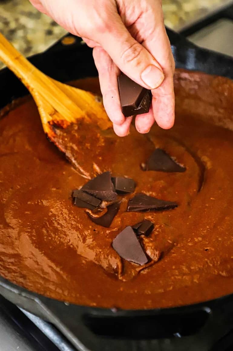 A person dropping chunks of dark chocolate into a skillet filled with a velvety mole sauce.
