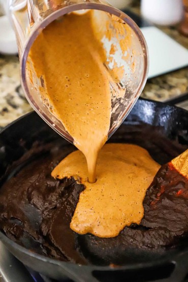 A beige creamy mixture being poured from a blender holder into a cast-iron skillet filled with a dark chili puree mixture.