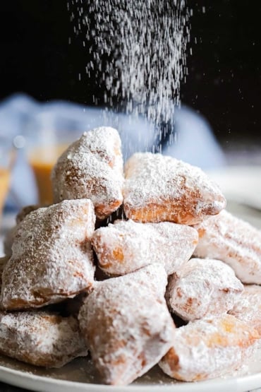 A circular plate filled with a pile of freshly fried beignets with powdered sugar being sprinkled over the top.