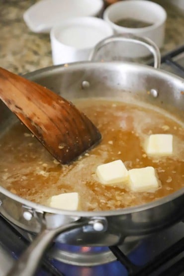 A person using a wooden flat spatula to stir small pallets of butter in a large skillet filled with simmering broth and minced garlic.