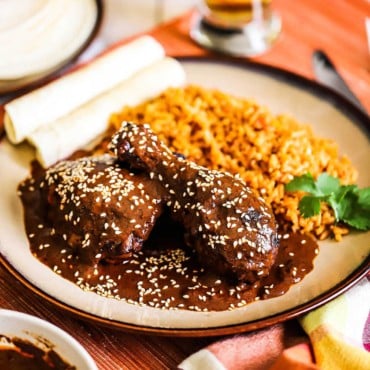 A front-view of a brown plate holding a serving of chicken mole next to Mexican rice, rolled tortillas, and a sprig of cilantro.