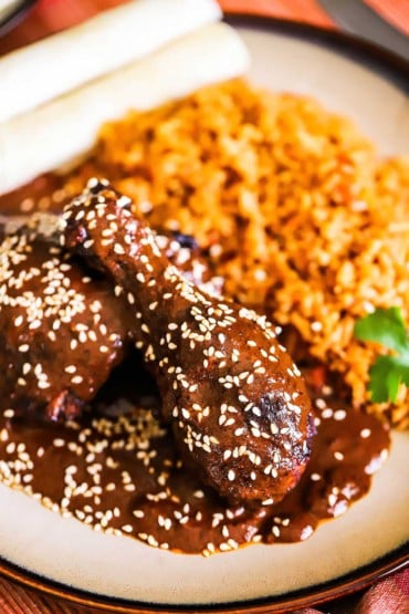 A close-up view of two pieces of chicken mole covered with sesame seeds and sitting next to a serving of Mexican rice all on a brown plate.