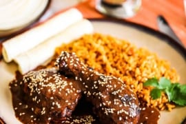 A front-view of a brown plate holding a serving of chicken mole next to Mexican rice, rolled tortillas, and a sprig of cilantro.