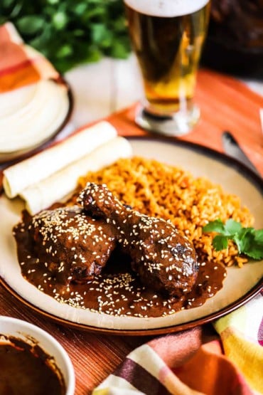 A front-view of a brown plate holding a serving of chicken mole next to Mexican rice, rolled tortillas, and a sprig of cilantro.