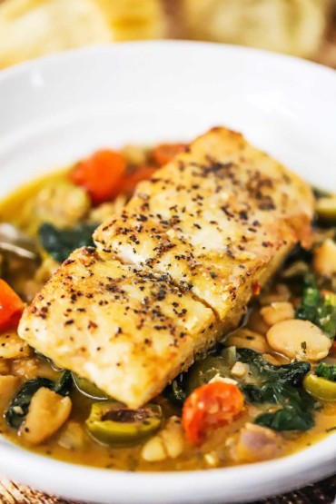 A close-up view of a seared fillet of halibut sitting on top of sautéed spinach, tomatoes, olive, and beans all in a savory sauce.