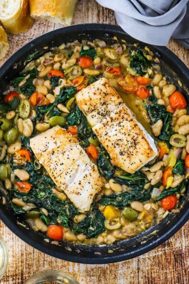 An overhead view of a large black skillet filled with sauteed vegetable in a white bean sauce with two halibut fillets nestled in the middle.