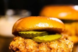 A close-up side view of a fried chicken sandwich with pickles and remoulade between toasted brioche buns.