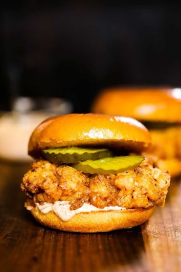 A close-up side view of a fried chicken sandwich with pickles and remoulade between toasted brioche buns.