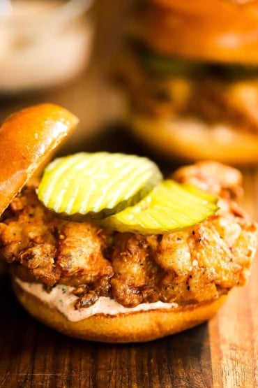 A fried chicken cutlet that it sitting on a toasted brioche bun and spicy remoulade with a couple of pickles on top.