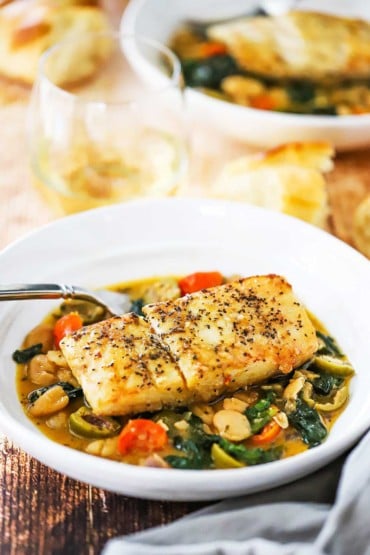 A side-view of a white pasta bowl filled sautéd vegetables and white bean in a broth and a fillet of halibut nestled on top.