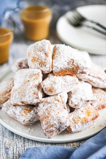 A tall pile of freshly fried beignets on a white circular plate next to a couple glass mugs of coffee.