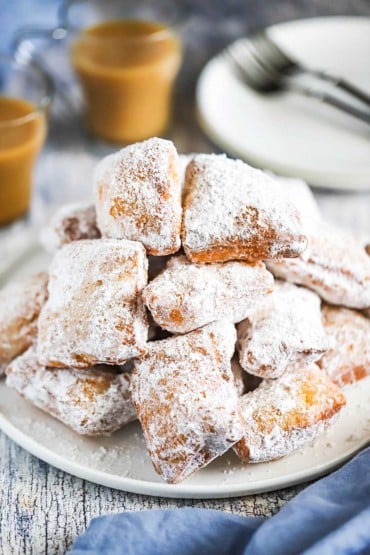 A large pile of freshly fried beignets on a white round plate with cups of coffee on the side.