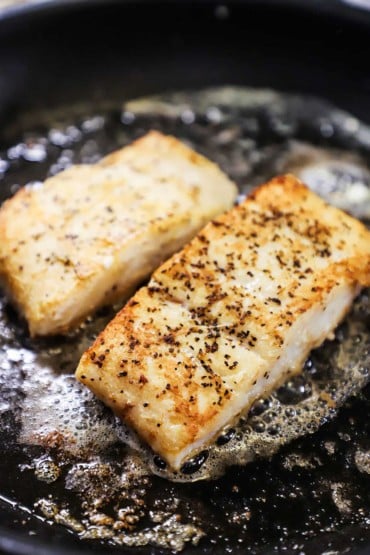 Two fillets of halibut that are seasoned with salt and pepper and are being seared in butter and olive oil in a large nonstick skillet.