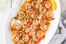 An overhead view of an oval platter filled with shrimp scampi being flanked by lemon wedges and a large golden serving spoon.